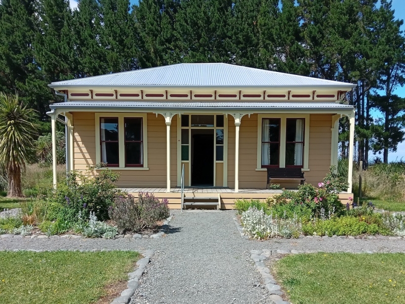 Photograph of the front of a tan-coloured colonial-era villa with verandah and corrugated iron roof. There is a gravel path that runs up to the front steps of the verandah and around two garden beds on either side. Behind the house is a wall of pine trees.