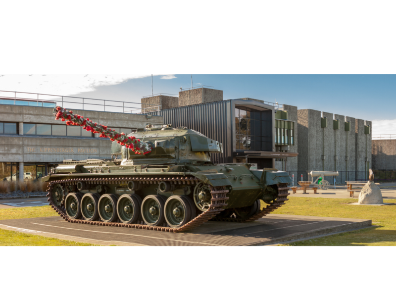 Photograph of World War 2-era tank with flowers wrapped around its main gun, situated on a platform in front of a brutalist-style building complex. The words "The Kippenberger Pavillion" appear in brass lettering on the front of the nearest building.