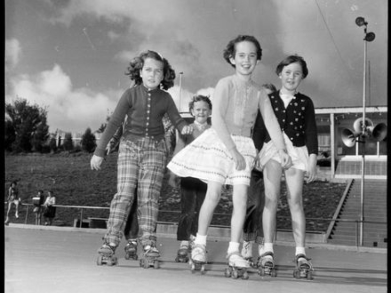 Black and white photograph taken from a low angle depicting five girls on roller skates. They are looking into camera as they appear to move forward, skating on a smooth concrete rink bordered by a fenced grassy slope, with steps leading up to a building at the top of the slope.