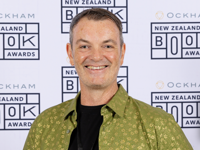 Head-and-shoulders photograph of a man smiling at camera, in front of a white wall with a repeating logo that reads: "Ockham New Zealand Book Awards".