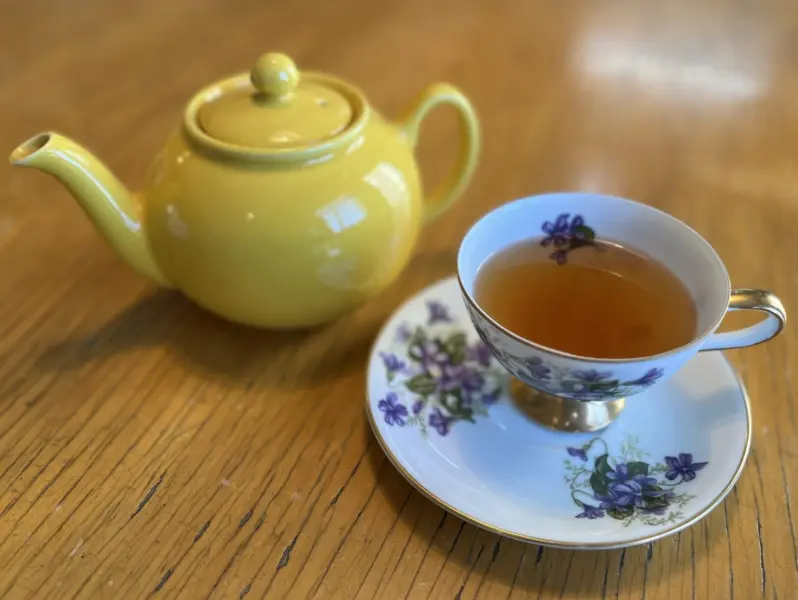 A white teacup and saucer with floral pattern beside a yellow teapot, on a wooden surface
